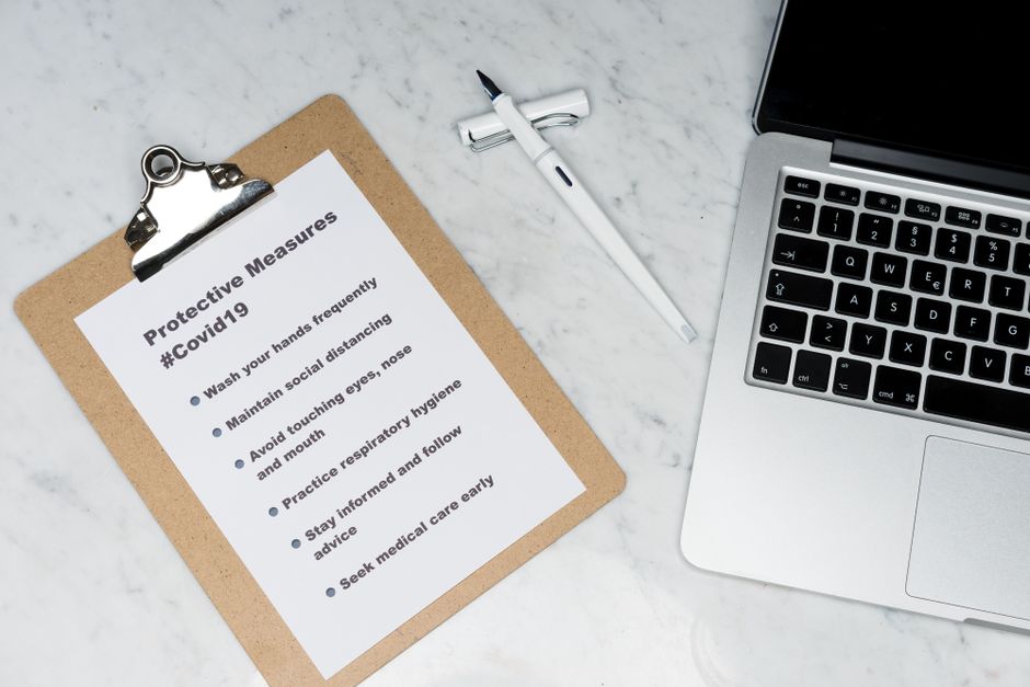 Flat lay of a clipboard listing Covid-19 safety measures beside a laptop on a marble surface.