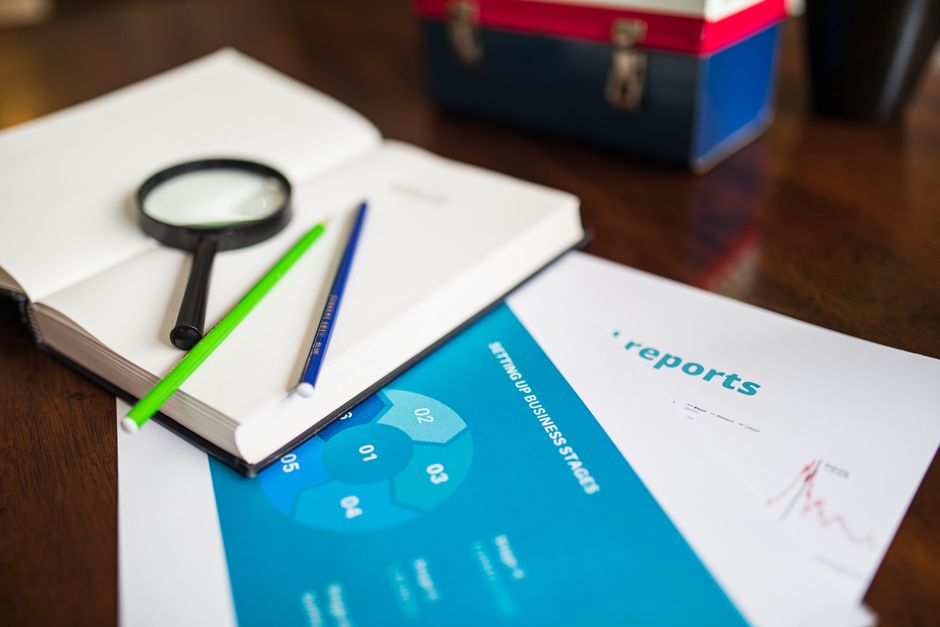 Open book with magnifying glass, pencils, and a business report on a wooden desk.